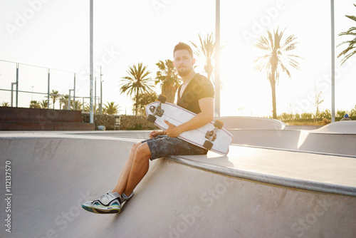 Young man with a skateboard is sitting in a scate park on a sunny day. A skater boy is looking aside while relaxing in a scate center. Flare light. photo