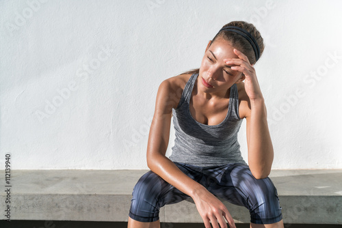 Tired fitness running woman sweating taking a break of cardio workout difficult training. Exhausted runner sitting with headache feeling exhaustion and dehydration from working out at outdoor gym.