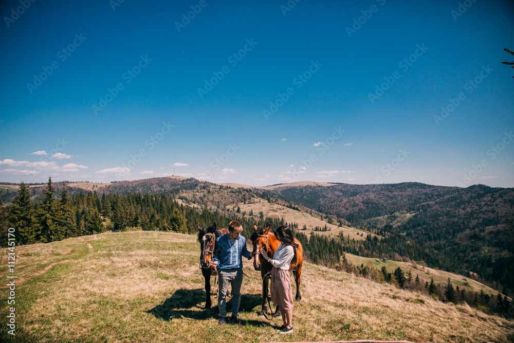 portrait of a cute couple with a horse in a mountains with blues sky. Couple walk and kiss near two horses. portrait of a cute couple with a horse in a mountains with blues sky. Couple walk and kiss near two horses.