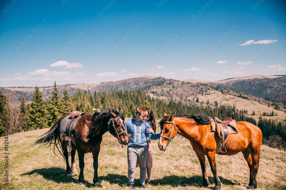 portrait of a cute couple with a horse in a mountains with blues sky. Couple walk and kiss near two horses. portrait of a cute couple with a horse in a mountains with blues sky. Couple walk and kiss near two horses.