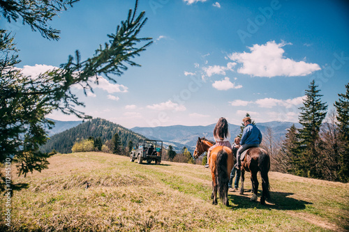 Young couple riding a brown horse at countryside at summer in the mountains with blue sky and pine tree. Woman in dress © nataliakabliuk