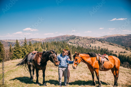 portrait of a cute couple with a horse in a mountains with blues sky. Couple walk and kiss near two horses.