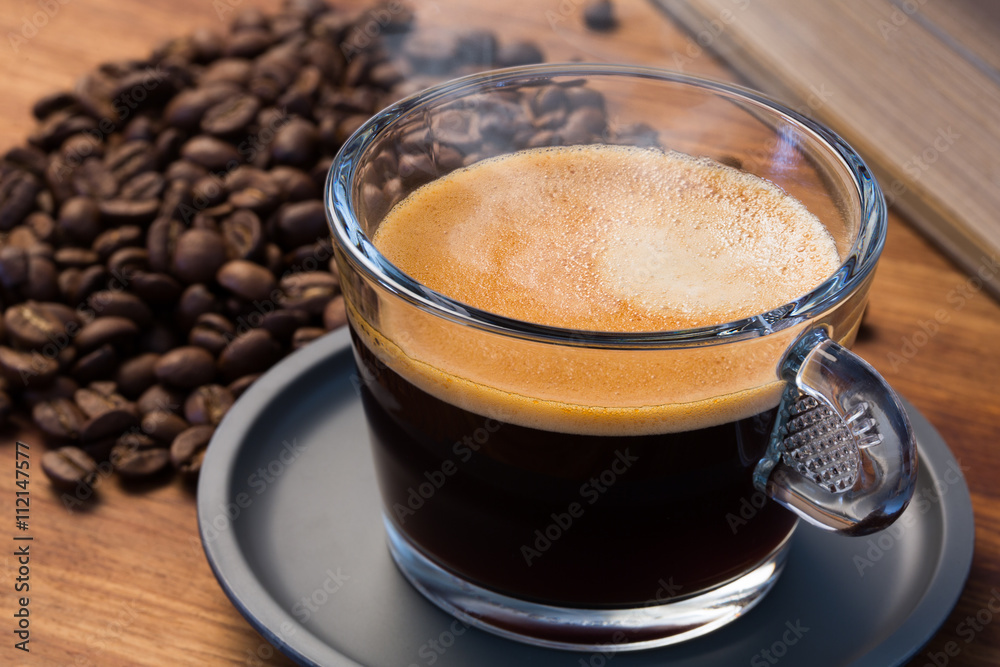 A transparent glass cup of hot steamy espresso on a saucer standing on a wooden table top, surrounded by coffee beans and a vintage book A transparent glass cup of hot steamy espresso on a saucer standing on a wooden table top, surrounded by coffee beans and a vintage book