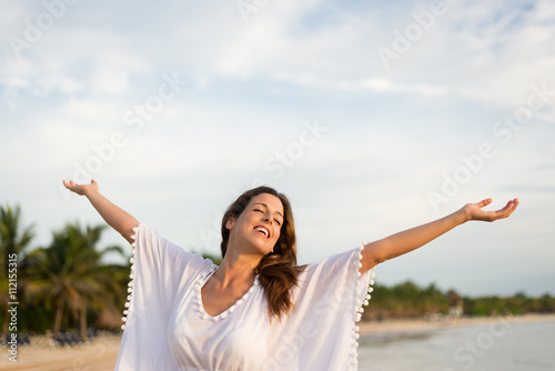 Blissful woman at tropical beach enjoying relax, freedom and vacation. Brunette female raising arms to the sky. Summer happiness and leisure.