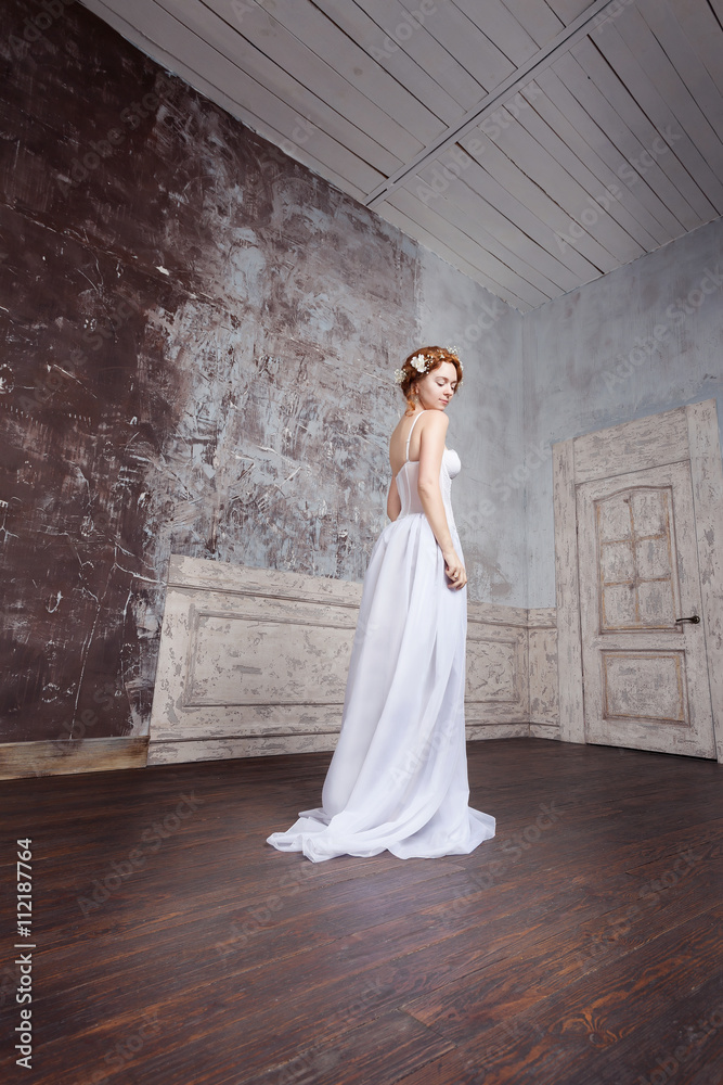 Beautiful young bride in a long wedding dress is standing in an empty room. White flowers in her red hair. She is pale, elate and tender. Beautiful young bride in a long wedding dress is standing in an empty room. White flowers in her red hair. She is pale, elate and tender.