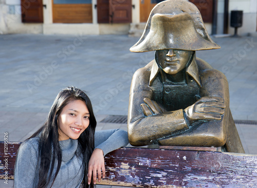 Canvas Print Asian tourist woman sitting on bench with Napoleon soldier at the Main square, B