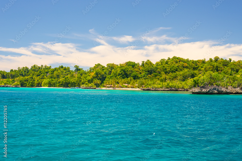 Rocky coastline of island spotted by islets and covered by dense lush green jungle in the colorful sea of the remote Togean Islands (or Togian Islands), Central Sulawesi, Indonesia.