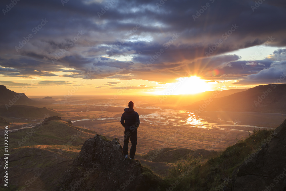 Silhouette of a man standing on a ledge of a mountain, enjoying the beautiful sunset over a  river valley in Iceland.