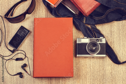 Composición con un libro de tapa naranja junto a una cámara de fotos, un bolso y gafas sobre un fondo de madera rústica. Vista superior y de cerca. Concepto estilo de vida