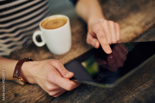 Close-up of woman hand pointing on screen of tablet pc. Young female browsing social network at wooden table with coffee cup. 