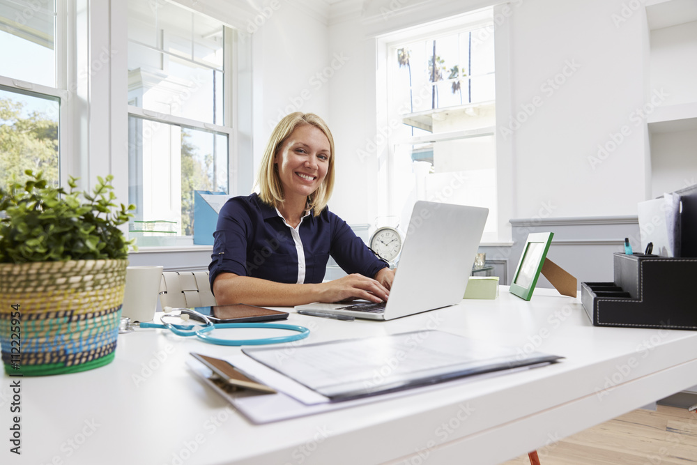 Fototapeta premium Female Doctor Sitting At Desk Working At Laptop In Office