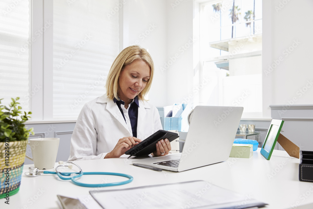 Female Doctor Sitting At Desk Using Digital Tablet In Office Female Doctor Sitting At Desk Using Digital Tablet In Office