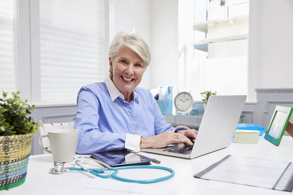 Female Doctor Sitting At Desk Working At Laptop In Office Female Doctor Sitting At Desk Working At Laptop In Office