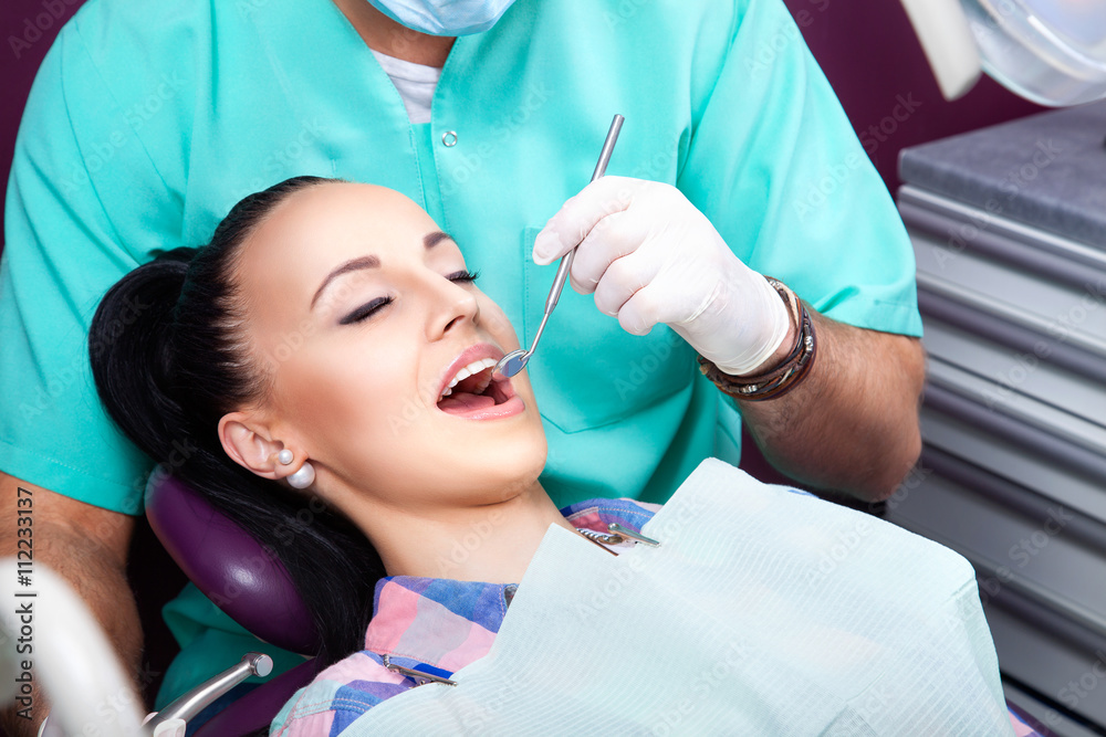 Fototapeta premium Young beautiful woman patient with white straight teeth and closed eyes sitting in dental chair while male doctor in white gloves examining her teeth with help of dental mirror. Healthcare, medicine.