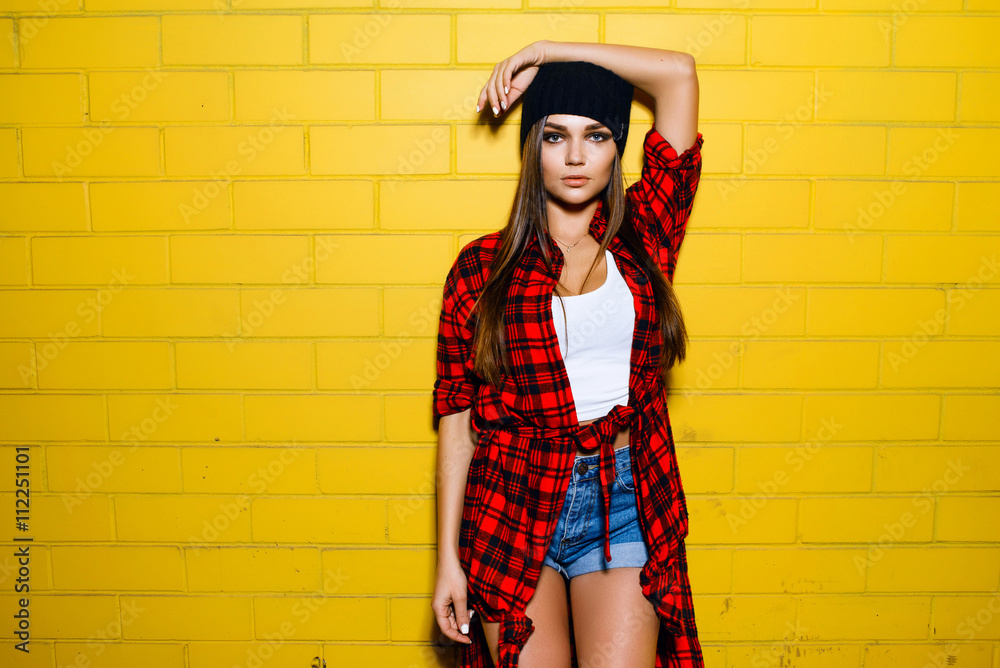 Beautiful young sexy hipster girl posing and smiling near urban yellow wall  background in red plaid shirt, shorts, hat. foto de Stock | Adobe Stock