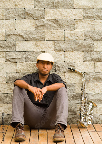 African man wearing sixpence hat and dark shirt sitting down leaning against wall, saxophone next to him photo