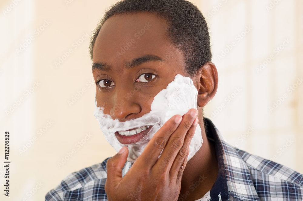 Young man wearing white blue square pattern shirt applying shaving foam onto face using hands, looking into camera Young man wearing white blue square pattern shirt applying shaving foam onto face using hands, looking into camera