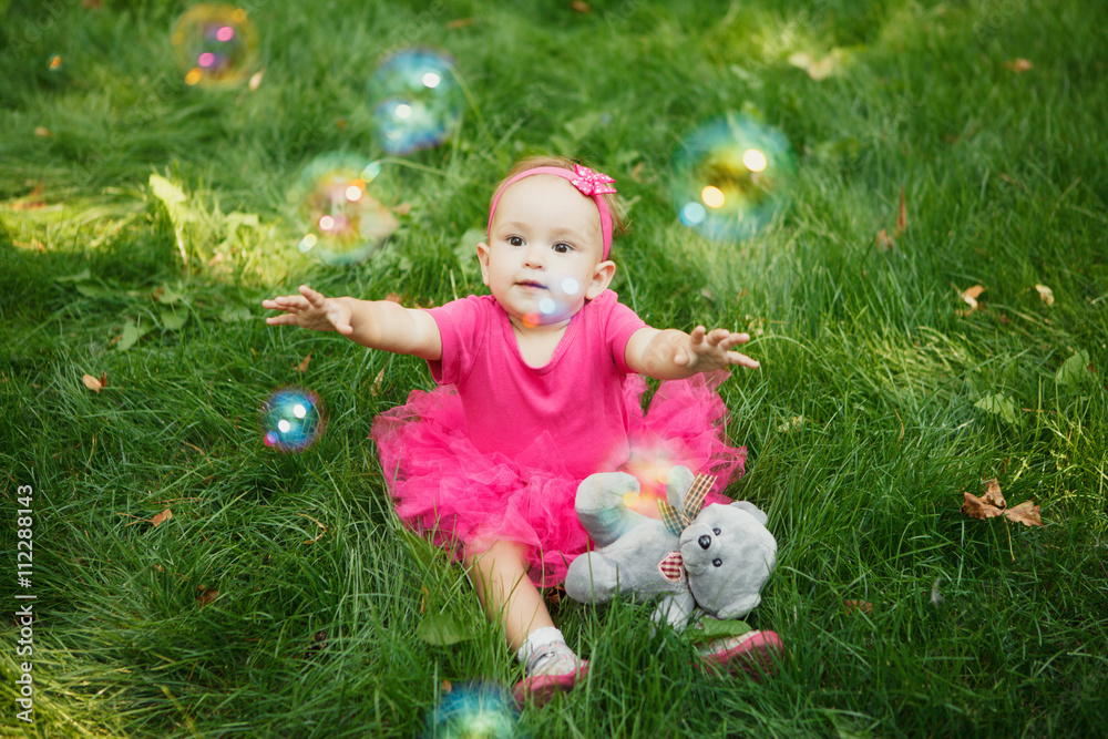 Baby girl sitting on grass outdoors dressed in pink dress with teddy bear  catching soap bubbles. Happy kid. Happy childhood. Stock Photo | Adobe Stock
