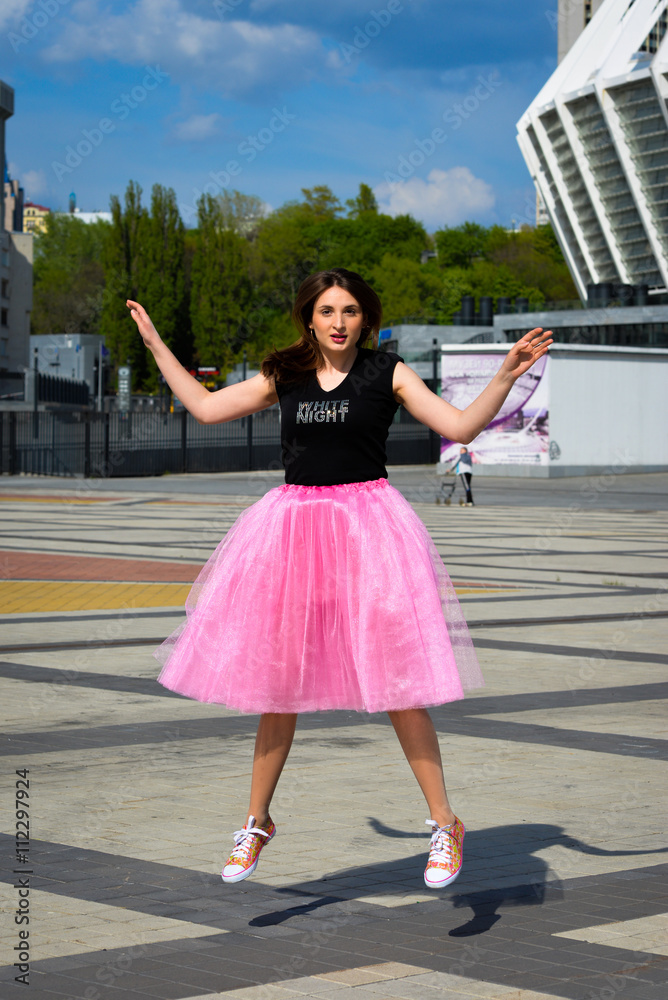 Beautiful sexy young hipster girl outdoors.Posh woman with long brunette hair evening makeup wearing light fluffy pink rosy skirt and vans sneakers posing outdoors in the city urban style in summer