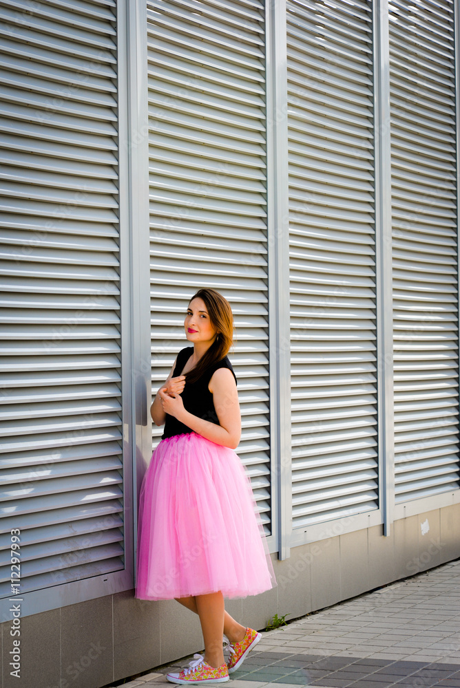 Fototapeta premium Beautiful sexy young hipster girl outdoors.Posh woman with long brunette hair evening makeup wearing light fluffy pink rosy skirt and vans sneakers posing outdoors in the city urban style in summer