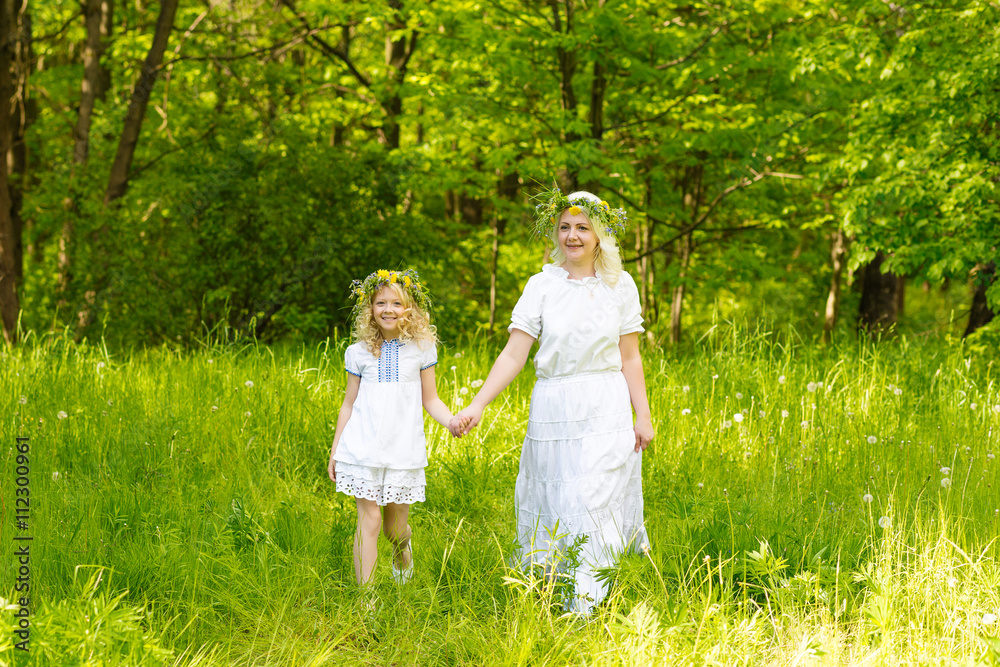 Fototapeta premium Beautiful family goes hand in hand. Happy family resting on the nature in the summer. Mom and daughter with wreath in the flowers in the park