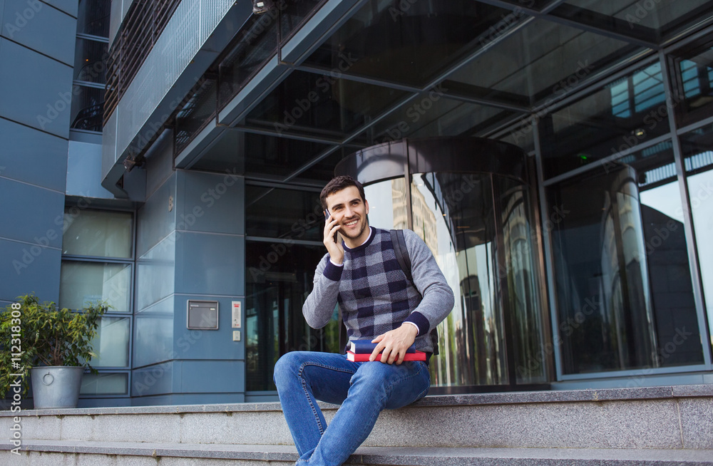 Smiling young man holding book and speaking on mobile phone in  city on university stair .Young smiling student  outdoors  with tablet and mobile phone.Life style.City