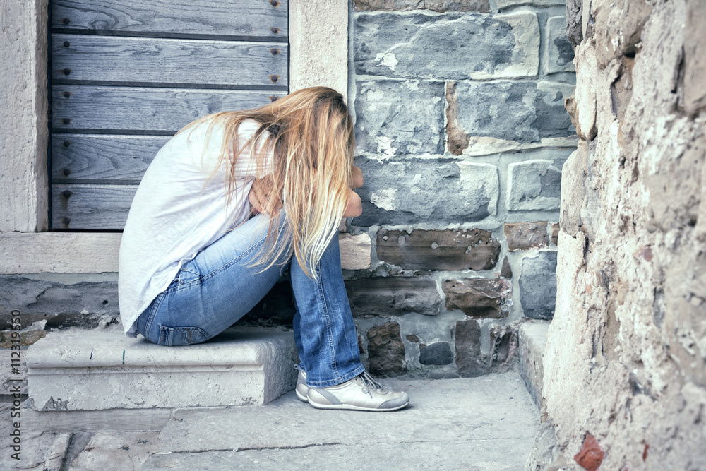 Sad and lonely blonde women sits in front of a stone aged house. Long hair heartbroken woman facing down alone. Sad and lonely blonde women sits in front of a stone aged house. Long hair heartbroken woman facing down alone.