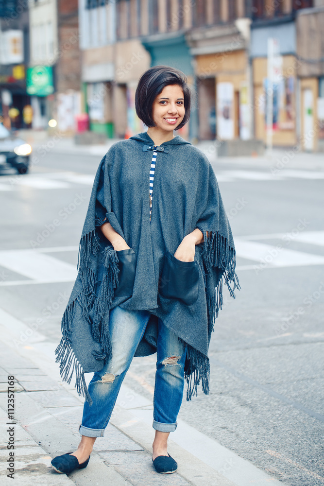 Portrait of beautiful smiling young hipster latin hispanic girl woman with  short hair bob, in torn jeans, grey blue poncho cape, standing outside in  city, looking in camera, street fashion concept Stock