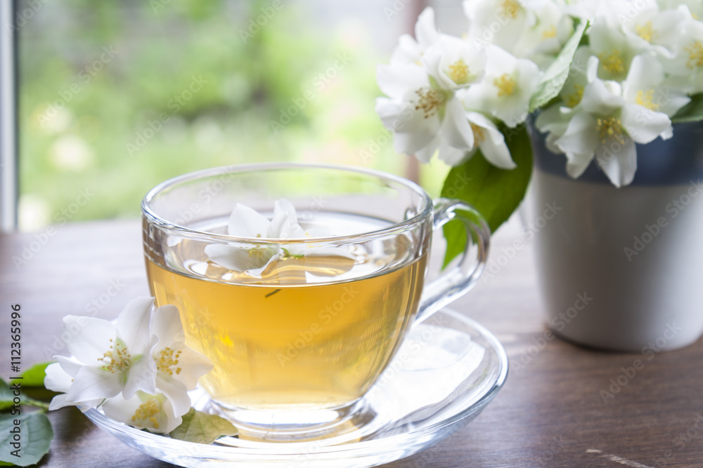 Transparent glass cup of tea with jasmine and blooming jasmine branch in blue ceramic cup on wooden table Transparent glass cup of tea with jasmine and blooming jasmine branch in blue ceramic cup on wooden table