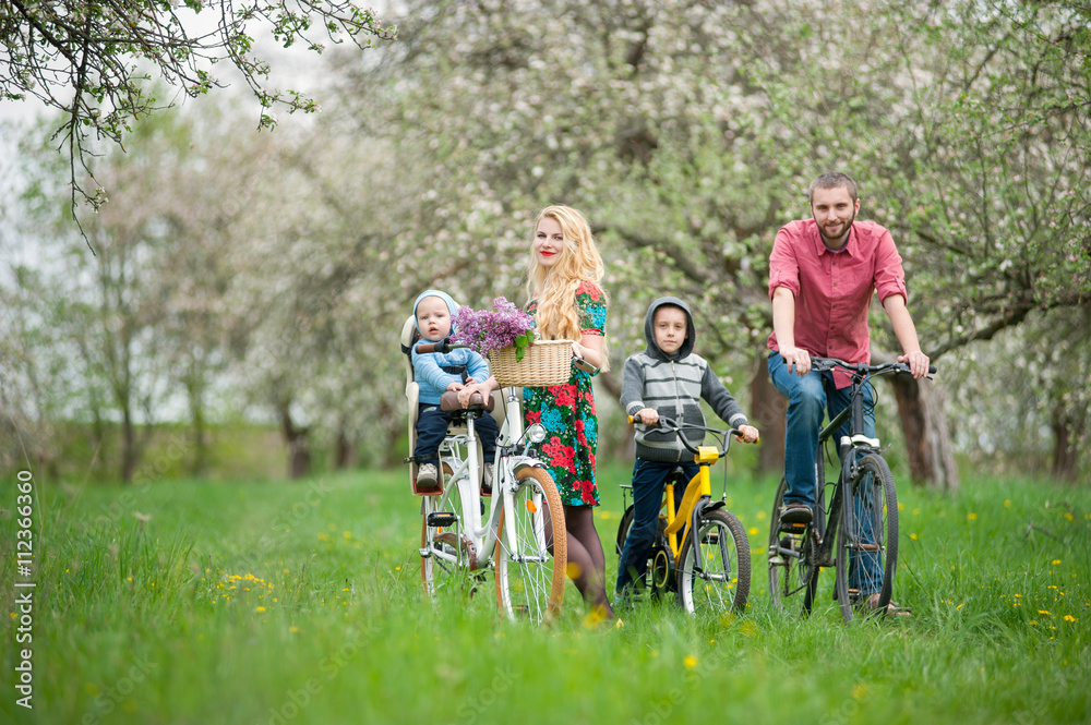 Happy family on a bicycles in the spring garden. Father and son on bikes, mom holding bike and baby sitting in bicycle chair, in the basket lay a bouquet of lilacs Happy family on a bicycles in the spring garden. Father and son on bikes, mom holding bike and baby sitting in bicycle chair, in the basket lay a bouquet of lilacs