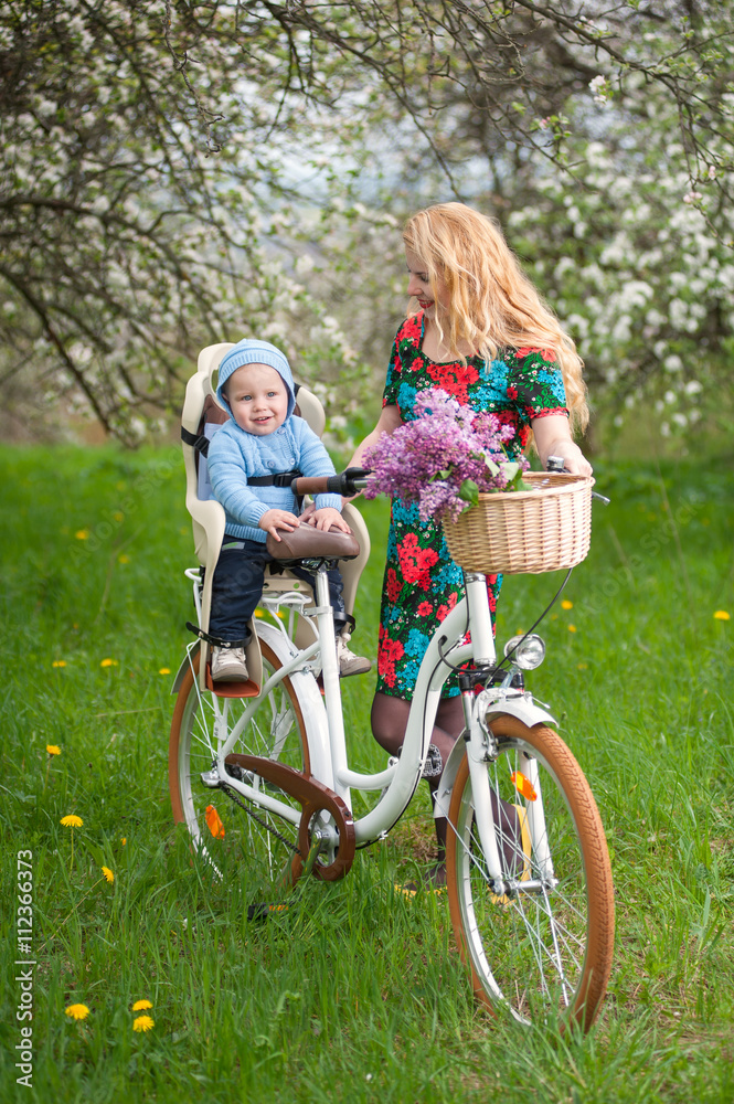 Blonde female with long hair and red lips in flowered dress holding baby in bicycle chair, in the basket lay a bouquet of lilacs, against the background of blooming trees