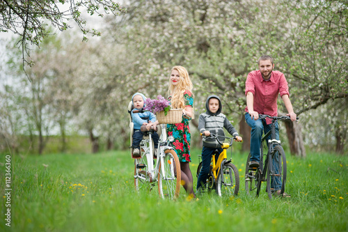 Happy family on a bicycles in the spring garden. Father and son on bikes, mom holding bike and baby sitting in bicycle chair, in the basket lay a bouquet of lilacs
