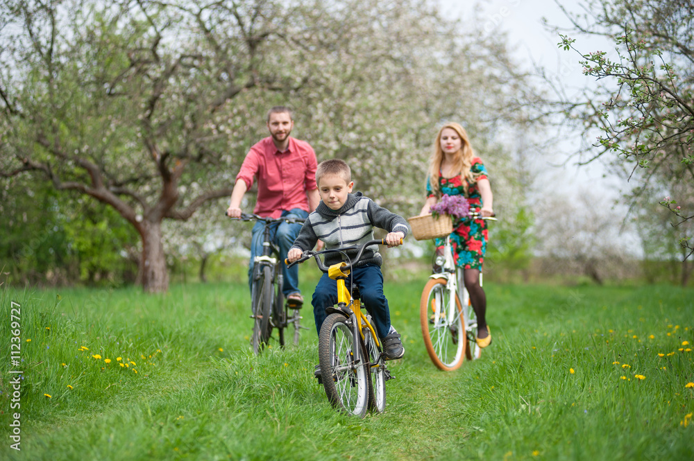 Happy boy on a bicycle in front of and behind its parent in the spring blooming garden riding bikes. Family having fun against the fresh greenery Happy boy on a bicycle in front of and behind its parent in the spring blooming garden riding bikes. Family having fun against the fresh greenery