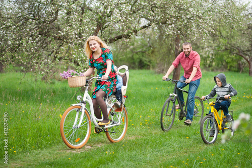 Young happy family with two kids riding bicycles in spring garden  little baby in bicycle chair  in the basket lay a bouquet of lilacs