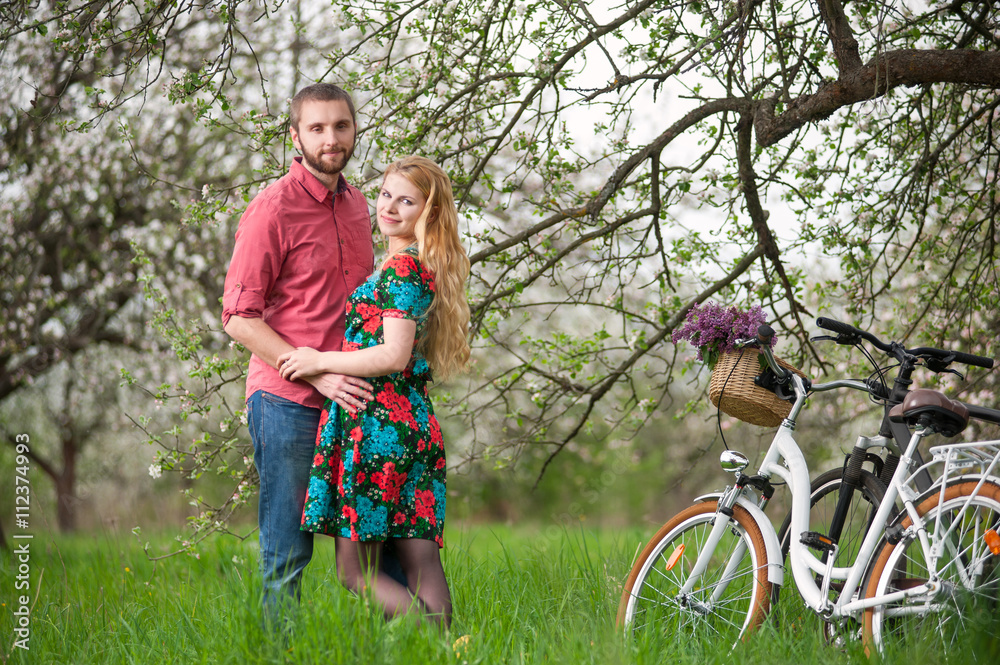 Romantic couple standing near the bicycles and tree, holding hands and looking to the camera in spring garden. Female with long blond hair wearing flowered dress and man in a red shirt and jeans