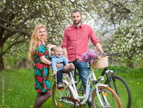 Happy family on a bicycles in the spring garden. Mother holding bike and baby sitting in bicycle chair. Against the background of blooming fresh greenery