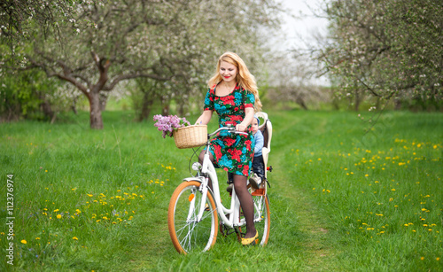 Happy mother with long blonde hair in dress riding city bicycle with baby in bicycle chair, in the basket lay a bouquet of lilacs, against the background of blooming fresh greenery in spring garden