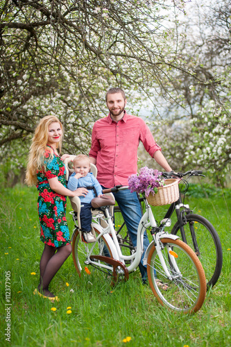 Beautiful family on a bicycles in the spring garden. Mother holding her bike and baby sitting in bicycle chair, against the background of blooming fresh greenery