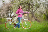 Smiling young woman wearing purple jacket and jeans with a vintage white bicycle and lilac flowers basket, looking to the camera in spring garden
