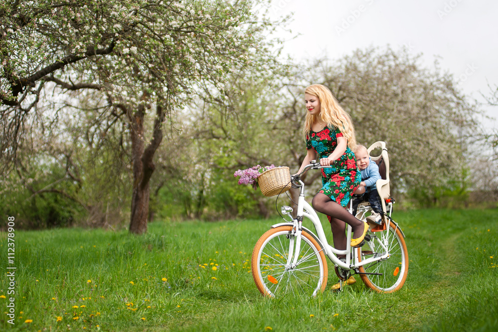 Young woman with long blonde hair in dress riding bicycle with baby in bicycle chair, in the basket lay a bouquet of lilacs, against the background of blooming fresh greenery in spring garden