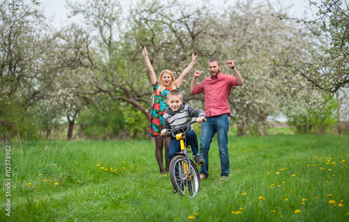 Happy child riding bicycle while his joyful parents celebrating success of his son. Family having fun against the blooming trees, dandelions and fresh greenery in spring garden