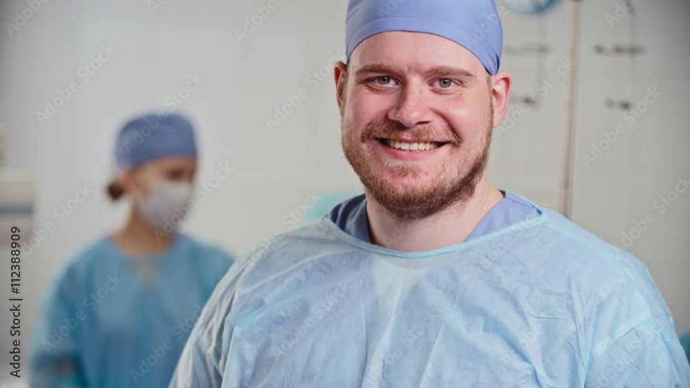 Young bearded surgeon turning to camera, taking off mask and smiling after surgical procedure in operating room, his colleagues working in the background, close-up shot on Sony NEX 700 + Odyssey 7Q