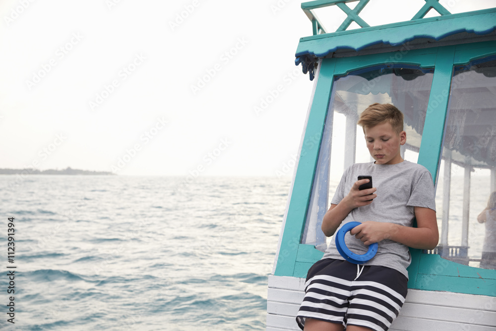 Teenage boy on boat using smartphone, ocean in the distance