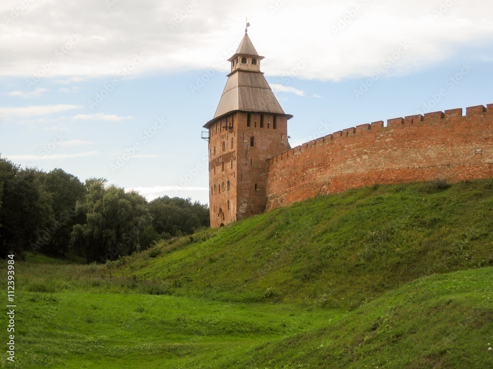 The tower and part of the walls of the ancient Kremlin in Veliky Novgorod, made of red bricks, located on a green hill.