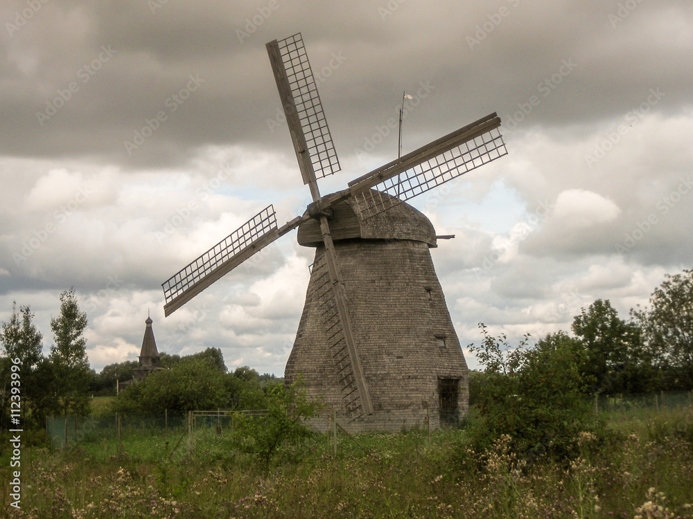 Old wooden mill among green grass and bushes in the summer. The sky with clouds.