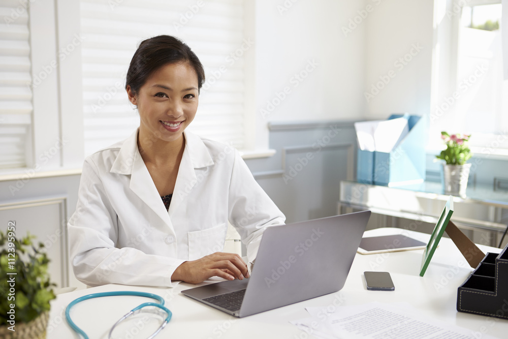 Female Doctor Sitting At Desk Working At Laptop In Office Female Doctor Sitting At Desk Working At Laptop In Office