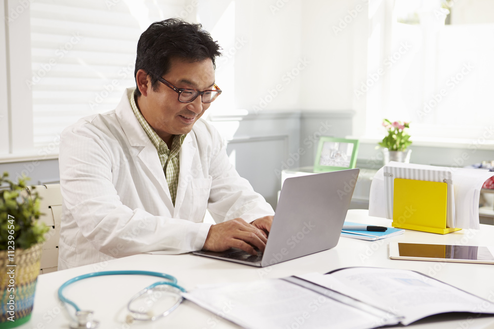 Male Doctor Sitting At Desk Working At Laptop In Office Male Doctor Sitting At Desk Working At Laptop In Office