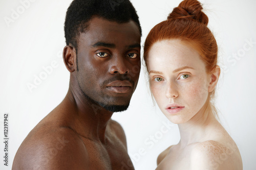Multicultural love and relationships concept: young nude redhead freckled Caucasian woman standing next to her shirtless African boyfriend, looking at the camera with serious face expression photo