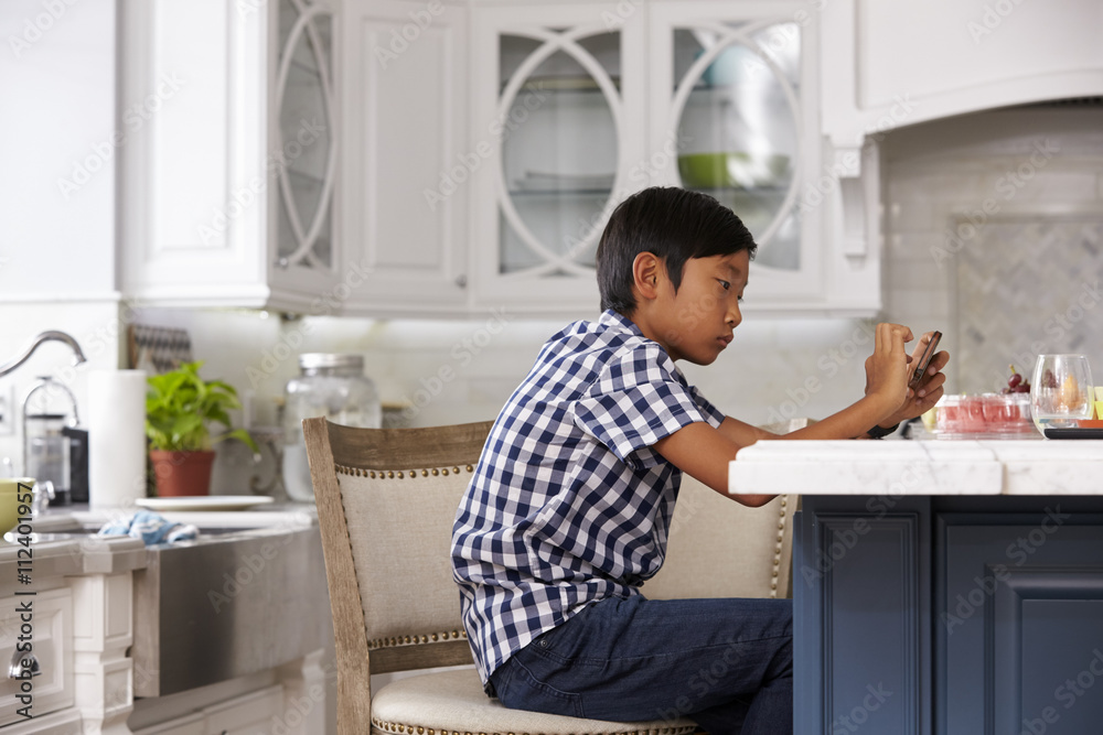 Young Asian Boy Playing Game On Mobile Device In Kitchen Young Asian Boy Playing Game On Mobile Device In Kitchen
