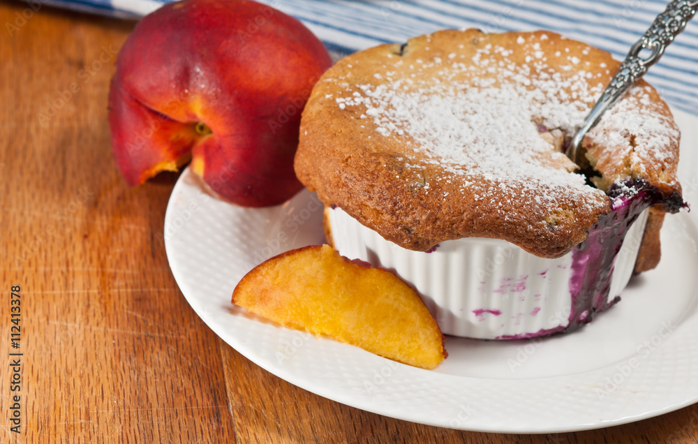 Blueberry Peach Cobbler in a ceramic cup on a white plate with sliced peach and a spoon Blueberry Peach Cobbler in a ceramic cup on a white plate with sliced peach and a spoon